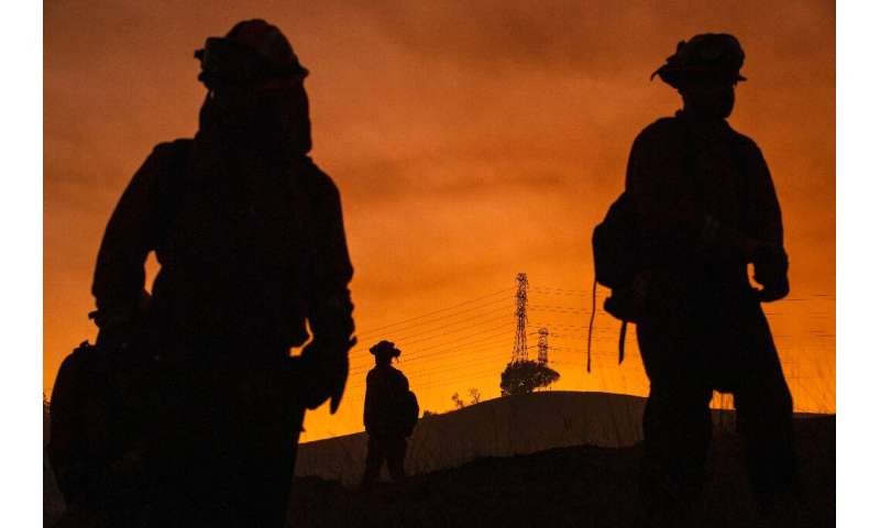 Members of an inmate firefighting crew are silhouetted against a hillside with a track of PG&amp;E lines during firefighting ope