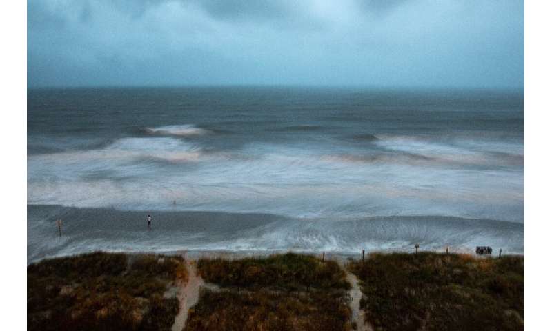 Large surf is swirled by the wind and captured by a long exposure, while a person stands on the shore as Hurricane Isaias approa