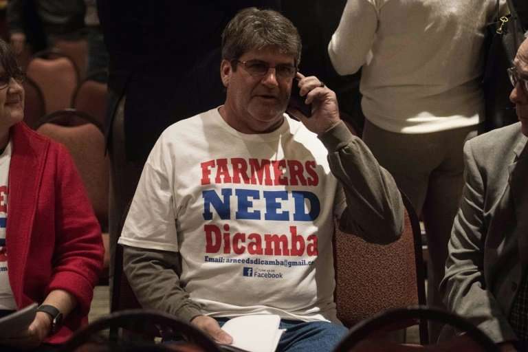 A farmer wearing a T-shirt reading &quot;Farmers Need Dicamba&quot; speaks on his phone during a break at a public hearing of th