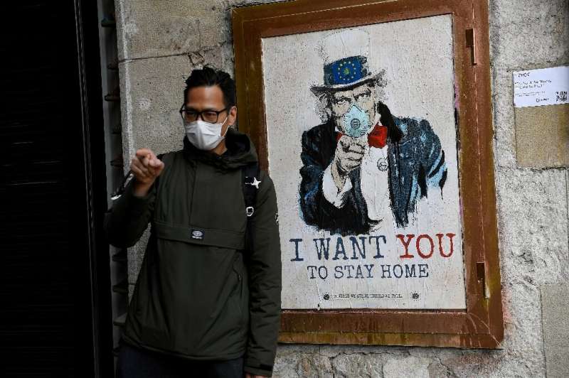 A man wearing a face mask poses with a poster by artist TVBoy in Barcelona, where authorities have ordered a widespread shop clo