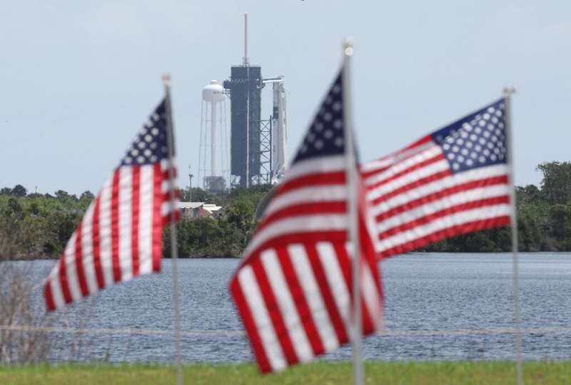 A SpaceX Falcon 9 rocket with the Crew Dragon spacecraft looms in the distance at launch complex 39A as American flags flutter i